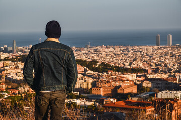 Man with black hat and denim jacket looking at Barcelona from the mountain in autumn