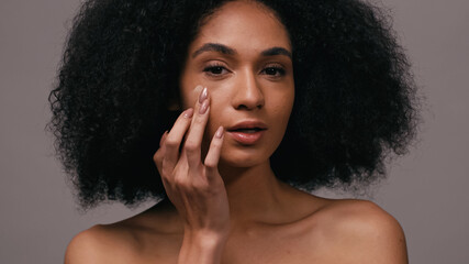 Fototapeta na wymiar curly african american woman applying face foundation with fingers isolated on grey