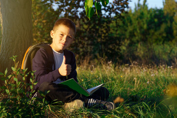 cute child reads a book under a tree. boy shows thumb up