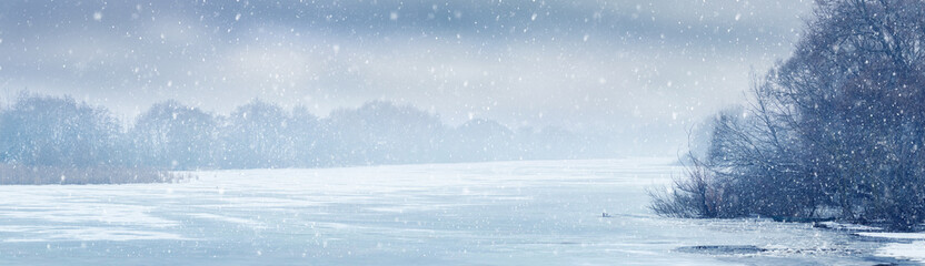 Winter view with ice and snow covered river and trees by the river during snowfall. Christmas background