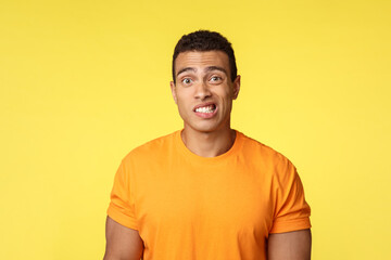 Yikes, awkward. Embarrassed young handsome man caught on lie, feel indesicive and slightly worried, cringe, smiling nervously and look camera with reluctance, stand yellow background - obrazy, fototapety, plakaty