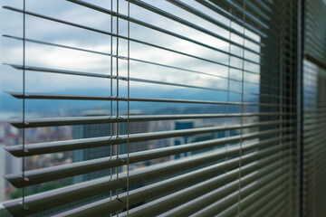 City houses, cloudy sky, mountains and sea are visible through the half-open roller blinds outside the window on a cloudy summer morning