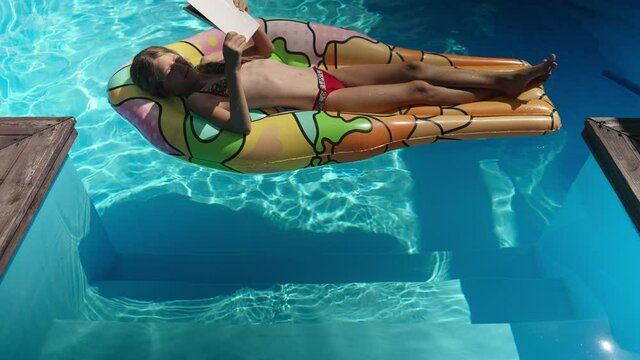 Happy smart girl sunbathing on inflatable mattress in swimming pool reading book in sunbeam. Wide shot of relaxed confident happy kid enjoying hobby on summer vacations tanning outdoors. Slow motion