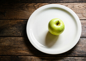 One green apple on a white ceramic plate, on a wooden table.