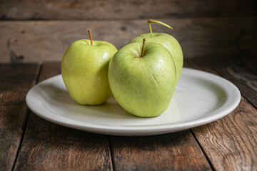 Three green apples on a white ceramic plate, on a wooden table.