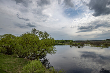 Landscape with a dramatic sky reflected in the river. Early spring, juicy May greens. Bright green foliage on trees and bushes.
