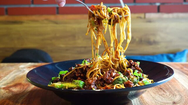 Woman with fork picking up indo chinese manchurian hakka street food noodles from a black bowl placed on a wooden table in a small street food eatery while travelling in Delhi