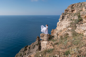 A beautiful young woman in a white light dress with long legs stands on the edge of a cliff above the sea waving a white long dress, against the background of the blue sky and the sea.