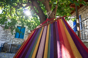 Swinging in colorful hammock with sun shining through green tree branches, summer recreation time in Greek traditional house yard with stone walls and blue shutters