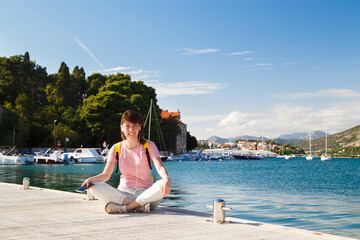 Smiling young female tourist sitting cross-legged on pier holding mobile phone with Croatian coastline in background. Bright sunny day in Dubrovnik, Croatia. Travel lifestyle concept.