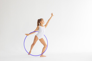 slim artistic teenager girl in white leotard trains on white background with hoop in her hands in...