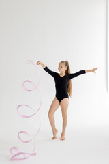 slim artistic teenager girl in black leotard trains on white background with ribbon in her hands in rhythmic gymnastic exercise, children's professional sports