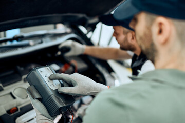 Close-up of auto mechanic uses diagnostic work tool while checking car battery with his coworker.