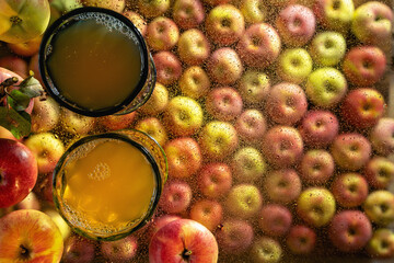 Two colored wine glasses filled with apple juice, surrounded and against the background of apples, stand on a transparent glass covered with water drops. Autumn still life, top view, selective focus