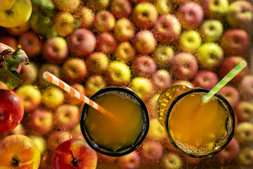 Two colored wine glasses filled with apple juice with paper straws, surrounded and against the background of apples, stand on a transparent glass covered with water drops. Autumn still life, top view