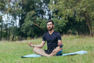 A young man with a beard sitting in the park on a mat meditates, performs exercises to improve breathing, active lifestyle