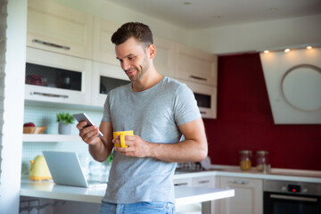 Handsome confident modern man in gray t-shirt stands with phone in hands and drinks coffee from the yellow cup on the kitchen at home