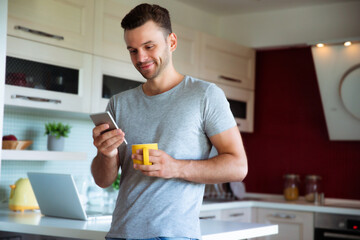 Handsome confident modern man in gray t-shirt stands with phone in hands and drinks coffee from the yellow cup on the kitchen at home © My Ocean studio
