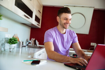 Happy handsome smiling young man in casual clothes is sitting while working on his laptop in the modern kitchen at home