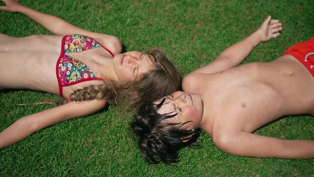 Top view of cute romantic couple of little kids lying on green summer grass warming in sunshine outdoors. Relaxed happy Caucasian boyfriend and girlfriend resting in sunlight on resort outdoors