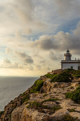 Fototapeta na wymiar Cap Blanc lighthouse on the rocky coast of the island of Mallorca. In the background s seascape of the Mediterranean Sea at sunset with clouds. Sublime landscape