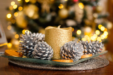 Silver color pine cones and golden candle in focus. Christmas tree with lights out of focus. Festive season concept. Decorating house for Christmas theme