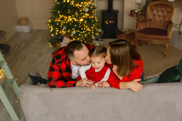 Parents kiss their daughter on Christmas and New Year's Eve against the background of a Christmas tree with lights and toys. A two-year-old girl grimaces and squints in response to a kiss. 