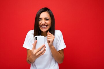 Photo shot of attractive happy joyful positive good looking young woman wearing casual stylish outfit poising isolated on background with empty space holding in hand and using mobile phone messaging