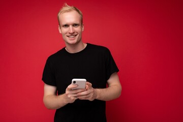 Photo of handsome smiling positive young blonde man isolated over red background wall wearing black t-shirt holding and using mobile phone writing sms looking at camera