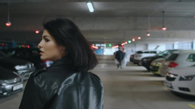 Following beautiful happy brunette woman wearing black leather jacket and sunglasses walking along underground parking with parked cars turns to camera flirts and smiling. Urban style fashion