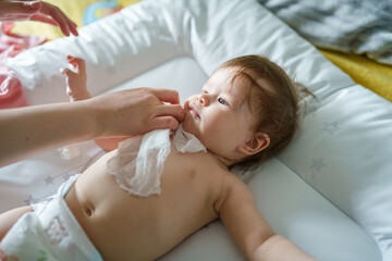 One small caucasian baby lying on the bed naked with hands of unknown woman mother using wet wipe...