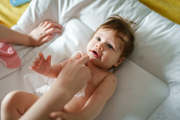 Obraz na płótnie Canvas One small caucasian baby lying on the bed naked with hands of unknown woman mother using wet wipe napkin towel to clean her skin growing up childhood hygiene care and parenthood concept