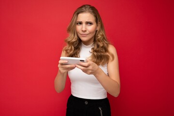 Naklejka premium Photo of concentrated beautiful young blonde woman wearing white t-shirt isolated on red background using smartphone and playing phone games looking to the side