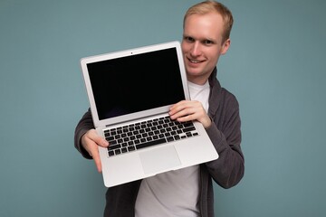 Close-up portrait of handsome smiling blonde man holding computer laptop with empty monitor screen with mock up and copy space wearing casual clothes looking at camera isolated over blue background