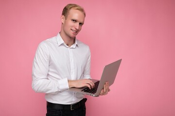 Side profile photo shot of handsome smiling confident blonde male person holding computer laptop typing on keyboard wearing white shirt looking at camera isolated over pink background