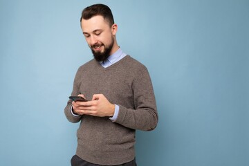 Photo shot of handsome positive good looking young man wearing casual stylish outfit poising isolated on background with empty space holding in hand and using mobile phone messaging sms looking at