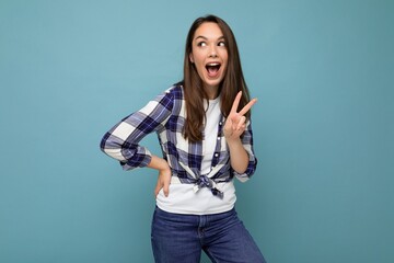Young positive happy amazed shocked beautiful winsom brunette woman with sincere emotions wearing check shirt poising isolated over blue background with copy space and showing peace sign