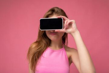 Photo of beautiful positive young blonde woman wearing pink top poising isolated on pink background with empty space holding in hand and showing mobile phone with empty display for mockup