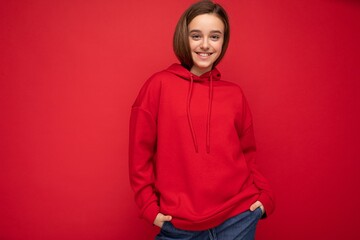 Photo shot of pretty cool happy smiling brunette little girl wearing trendy red hoodie standing isolated over red background wall looking at camera
