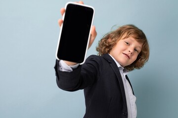 Handsome positive smiling boy with curly hair wearing suit holding phone isolated over blue...