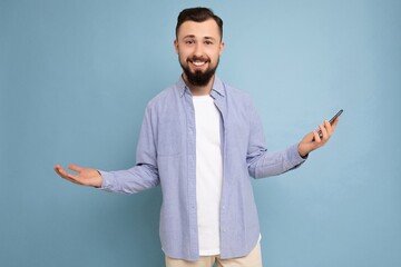 Photo of joyful smiling handsome young brunette unshaven male person with beard wearing stylish white t-shirt and blue shirt isolated over blue background with empty space holding in hand and using