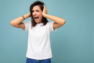 Portrait of emotional nervous young pretty nice brunette woman with sincere emotions wearing casual white t-shirt for mockup isolated on blue background with copy space and covering ears with hands