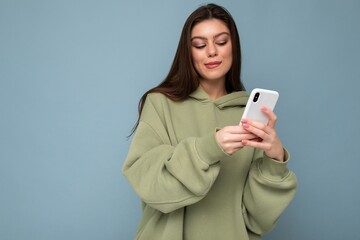 Fototapeta premium Sexy Beautiful young brunette woman wearing stylish green hoodie using mobile phone writing sms isolated on background looking at smartphone screen