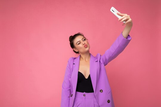 Photo of pretty young woman wearing purple suit taking selfie photo on the mobile phone isolated over pink background looking at phone camera