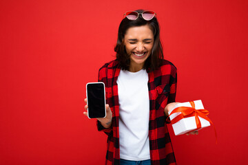 Laughing beautiful positive young brunette woman isolated over red background wall wearing white casual t-shirt and red and black shirt holding white gift box with red ribbon and mobile phone with