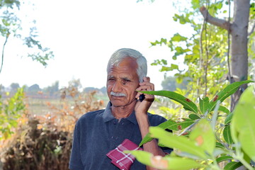 An elderly Indian farmer or gardener standing in a garden or field talking to an agricultural co-operator or specialist on a keypad mobile