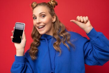 Naklejka premium Photo of beautiful smiling young woman good looking wearing casual stylish outfit standing isolated on background with copy space holding smartphone showing phone in hand with empty screen display for