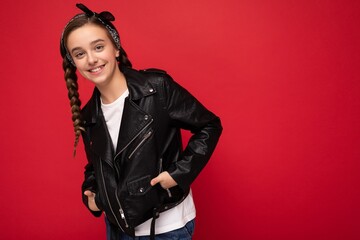 Shot of attractive positive smiling brunette little female teenager with pigtails wearing stylish black leather jacket and white t-shirt isolated over red background wall looking at camera