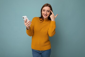 Photo of beautiful positive amazed young woman wearing orange sweater poising isolated on blue background with empty space holding in hand and using mobile phone with communicating with friends