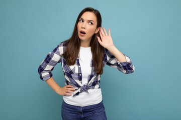 Young concentrated beautiful brunet woman with sincere emotions wearing trendy check shirt standing isolated on blue background with empty space and listening to something by putting hand on the ear